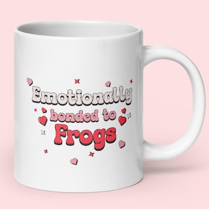 Puede incluir: Taza de cerámica blanca con el texto "Emotionally bonded to Frogs" en una fuente retro, decorada con corazones y estrellas rojas. La taza tiene un asa curva y está sobre un fondo rosa claro.