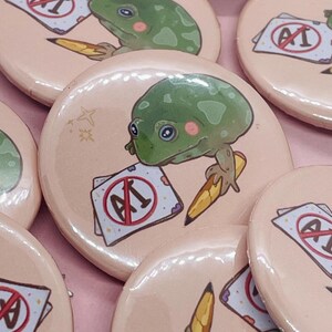 May include: A set of four round pink button pins featuring a green frog holding a pencil and a paper with a red circle and the text "AI" with a diagonal line through it. The frog has large eyes and a pink blush on its cheeks.