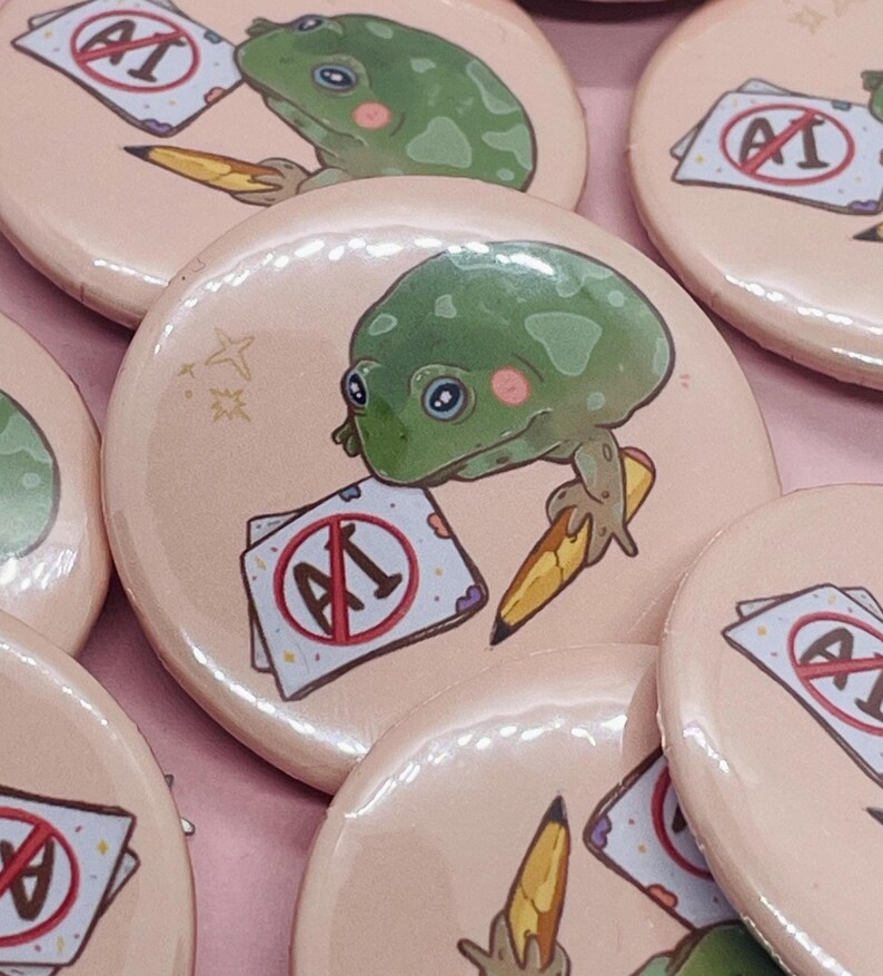 May include: A set of four round pink button pins featuring a green frog holding a pencil and a paper with a red circle and the text "AI" with a diagonal line through it. The frog has large eyes and a pink blush on its cheeks.