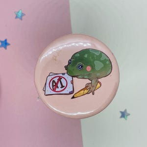 May include: A pink button with a green frog holding a pencil and a paper with a red circle and the text "A1" inside. The frog has a pink spot on its face.