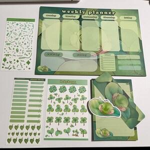May include: A set of green and white planner stickers featuring a weekly planner, planner blocks, lucky leaves, and plan & sparkle stickers. The weekly planner has sections for Monday through Sunday, with a reminder section. The planner blocks include various shapes and sizes of green rectangles, stars, and hearts. The lucky leaves sticker sheet features multiple green four-leaf clovers. The plan & sparkle sticker sheet features green swirls, stars, and other shapes.