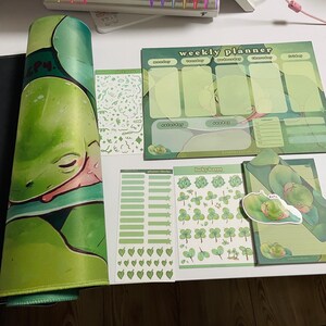 May include: A green frog themed weekly planner set with stickers. The set includes a planner, a sheet of planner blocks, a sheet of lucky leaves, and a sheet of hearts. The planner has a green frog design and the text "weekly planner" on the top. The stickers are all green and feature frogs, leaves, and hearts.