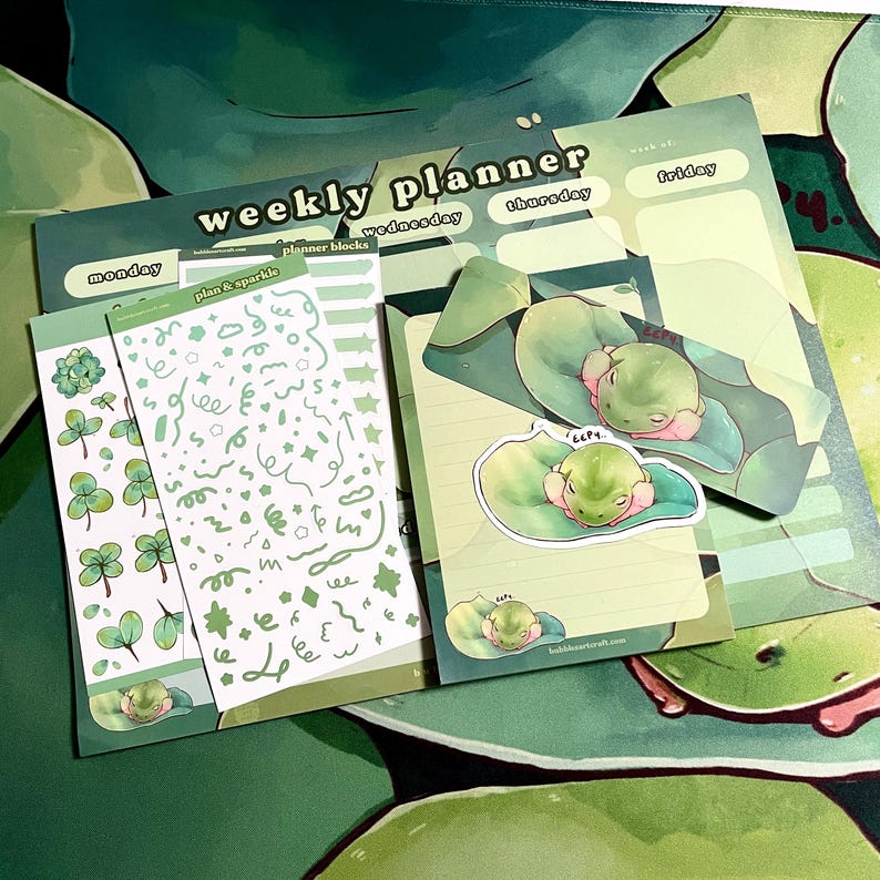 May include: A weekly planner with a green and white color scheme. The planner has a frog illustration on the cover and includes a sheet of stickers with frog illustrations and a sheet of stickers with green and white swirls, stars, and other shapes.