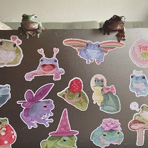 May include: A laptop computer decorated with frog stickers. The stickers feature frogs in various poses and colors, including green, blue, purple, and pink. Some stickers have text, such as "Frog" and "Forg".