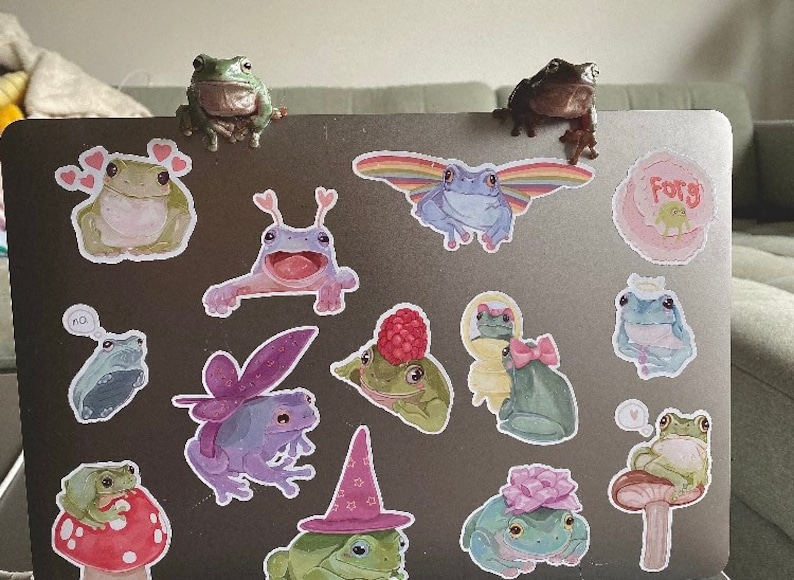 May include: A laptop computer decorated with frog stickers. The stickers feature frogs in various poses and colors, including green, blue, purple, and pink. Some stickers have text, such as "Frog" and "Forg".