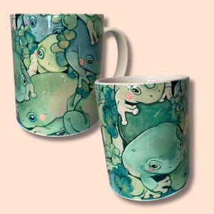 Könnte beinhalten: Eine weiße Keramiktasse mit einem grünen und blauen Design, das ein Muster aus Fröschen zeigt. Die Frösche sind in verschiedenen Posen dargestellt, einige mit geschlossenen Augen und andere mit offenem Mund. Die Tasse hat einen Henkel auf der rechten Seite.