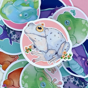 May include: A collection of frog stickers with various designs. The stickers feature frogs in different poses and colors, including green, blue, and white. Some stickers have floral accents.