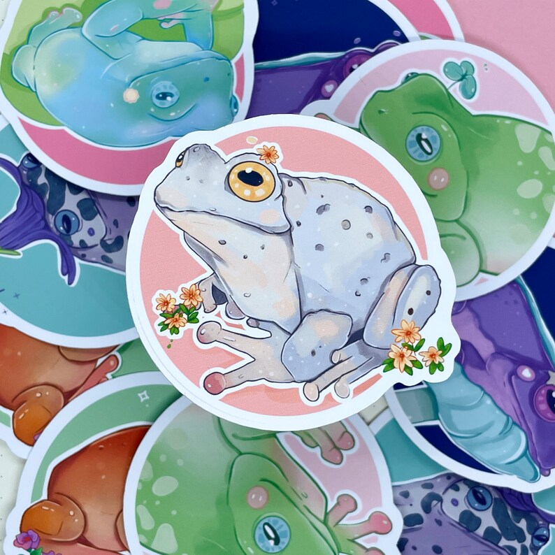 May include: A collection of frog stickers with various designs. The stickers feature frogs in different poses and colors, including green, blue, and white. Some stickers have floral accents.