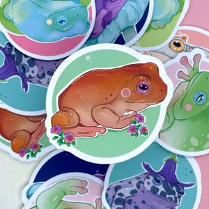 May include: A set of colorful frog stickers with different designs and poses. The stickers feature frogs with various colors, including green, blue, brown, and white. Some frogs have flowers and other details.