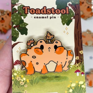 Puede incluir: Un pin de esmalte naranja y crema con una rana con acentos de hongos. El pin está etiquetado "Toadstool - enamel pin -" en una fuente caprichosa, con un fondo temático de bosque. El pin mide aproximadamente 3,8 cm de ancho.