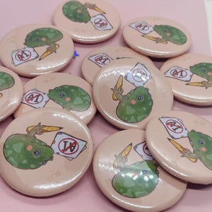 May include: A set of 10 round, pink button pins featuring a cartoon frog holding a pencil and a paper with a red circle and the letter 'F' inside. The frog is green with white eyes and a pink tongue. The background of the pins is a light pink with white stars.