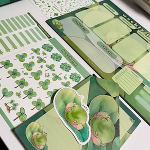 May include: A set of green and white planner stickers featuring a weekly planner, a sheet of clover stickers, and a sticker sheet with a frog on a leaf. The planner has sections for each day of the week, a reminder section, and the text "weekly planner". The clover stickers are in various sizes and shapes. The frog sticker has the text "eep!" on it.