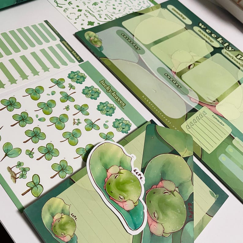 May include: A set of green and white planner stickers featuring a weekly planner, a sheet of clover stickers, and a sticker sheet with a frog on a leaf. The planner has sections for each day of the week, a reminder section, and the text "weekly planner". The clover stickers are in various sizes and shapes. The frog sticker has the text "eep!" on it.
