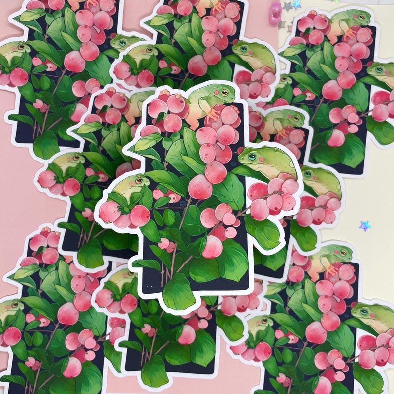 Puede incluir: Una colecci&oacute;n de pegatinas con un dise&ntilde;o caprichoso. Cada pegatina muestra una rana verde posada en una rama con bayas rosas y hojas verdes. Las pegatinas tienen un borde blanco y un fondo oscuro.