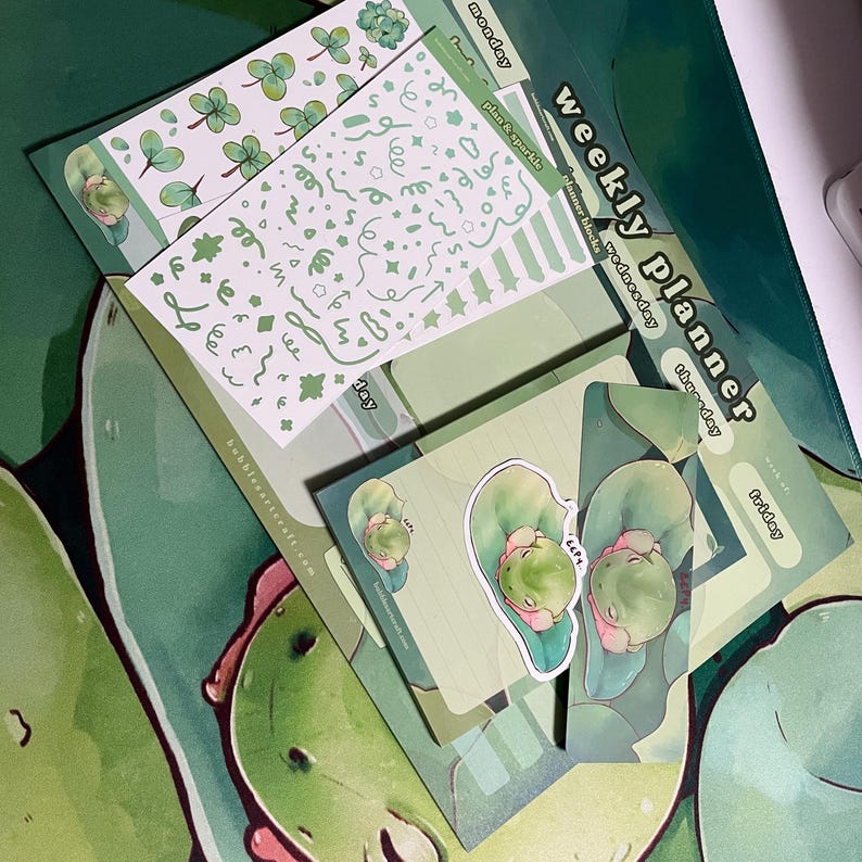May include: A green weekly planner with a frog illustration. The planner has sections for each day of the week, including Monday, Tuesday, Wednesday, Thursday, Friday, Saturday, and Sunday. The planner also includes a section for "mood", "plan & sparkle", and "hammer blocks".