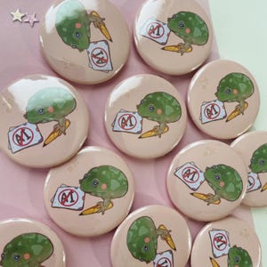 May include: A set of 10 round pink button pins featuring a cartoon frog holding a pencil and a paper with a red circle and the letter "A" with a line through it. The frog is green with a white belly and black eyes.