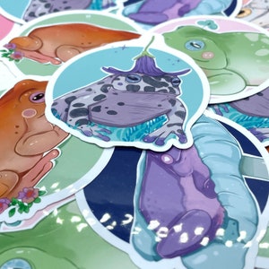 May include: A collection of frog stickers with various designs. The stickers feature frogs in different poses and colors, including a purple frog with a flower on its head, a green frog with a white belly, and a brown frog with a white spot on its back.