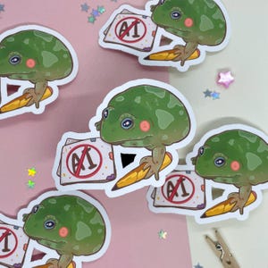 May include: Five stickers featuring a green frog holding a pencil and a paper with a red circle and the text "AI" with a diagonal line through it. The frog is holding a pencil and the paper is on a pink background.