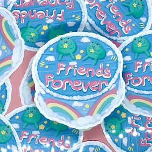 Könnte beinhalten: Ein blauer und rosa Aufkleber mit einem Kuchen-Design und zwei grünen Fröschen. Der Kuchen hat einen Regenbogen und Wolken. Der Text auf dem Kuchen lautet "Friends Forever".