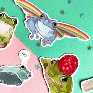 May include: Three frog stickers with different designs. One frog has rainbow wings, another has a raspberry on its head, and the third frog has a speech bubble that says "no".