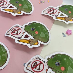 May include: Four stickers featuring a green frog holding a pencil and a paper with a red circle and the text "AI" with a diagonal line through it. The frog is holding a pencil in its right hand and the paper in its left hand. The stickers are on a pink and white background.