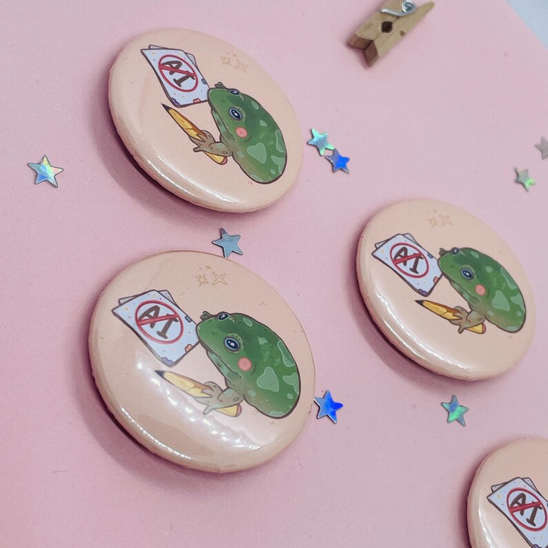 May include: Four round pink buttons with a green frog holding a pencil and a paper with a red circle and the letter A with a line through it. The frog is looking at the paper. The buttons are on a pink background with silver glitter.