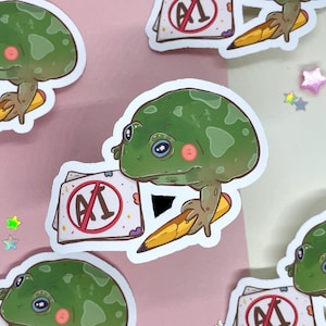 May include: A green frog with a red circle and a diagonal line through it, with the letter "A" inside the circle. The frog is holding a yellow pencil and is sitting on a white sticker with a pink and white background.