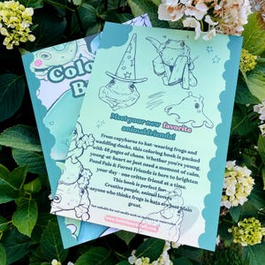 May include: Two coloring books are displayed, one partially visible. The visible book features a frog illustration with a wizard hat and the text "Meet your new favorite animal friends!". The book's cover is light green with a cartoon frog. The background is a mix of green foliage and white flowers.