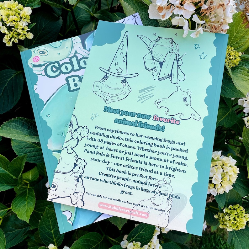 May include: Two coloring books are displayed, one partially visible. The visible book features a frog illustration with a wizard hat and the text "Meet your new favorite animal friends!". The book's cover is light green with a cartoon frog. The background is a mix of green foliage and white flowers.