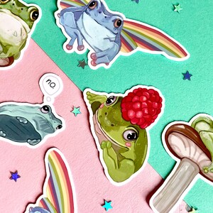 May include: A set of five frog stickers with different designs. The stickers feature frogs in various poses, including a frog wearing a rainbow, a frog with a raspberry on its head, and a frog with a thought bubble that says "no".