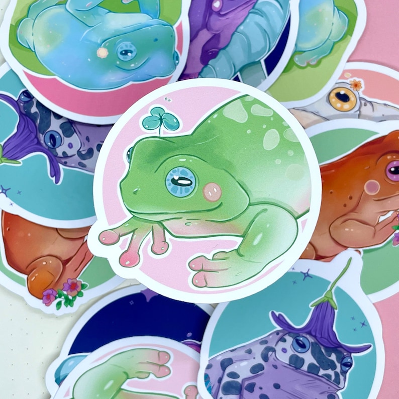 May include: A set of colorful frog stickers with different designs and poses. The stickers feature frogs with green, blue, purple, and orange colors. Some stickers have a pink background with a clover leaf design.