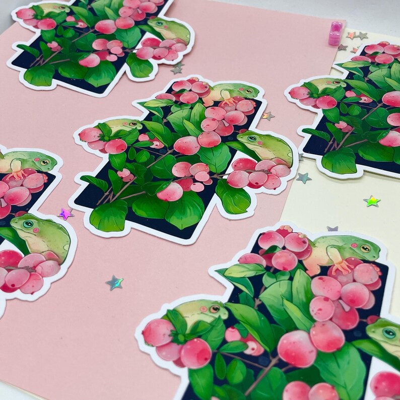 Puede incluir: Conjunto de pegatinas con ranas verdes posadas en ramas con bayas rosas y hojas verdes. Las pegatinas tienen un borde negro y est&aacute;n dispuestas sobre un fondo rosa y crema con confeti en forma de estrella.