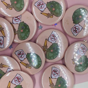 May include: A set of 10 round, pink button pins featuring a green frog holding a piece of paper with a red circle and the text "A1" crossed out. The frog is holding a pencil in its other hand.