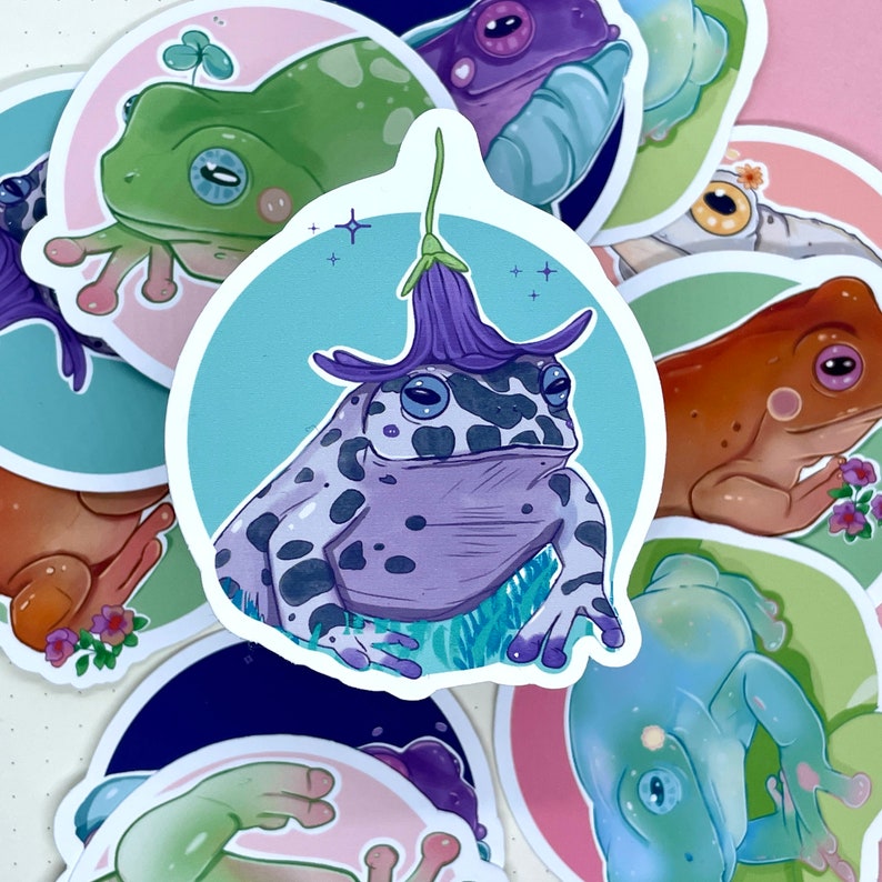 May include: A set of colorful stickers featuring various frog illustrations. The stickers feature frogs with different colors, patterns, and poses, including a frog wearing a purple flower on its head.