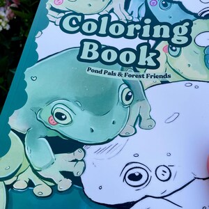 May include: A coloring book titled "Coloring Book" with the subtitle "Pond Pals & Forest Friends." The cover features a cartoon frog in shades of green and white. The frog has large eyes and a pink tongue. The book is open, revealing a partially colored page.