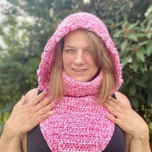 Può includere: Una sciarpa con cappuccio all'uncinetto rosa e bianca con collo a lupetto. La sciarpa è realizzata con un filato morbido e ha un motivo a punto testurizzato.