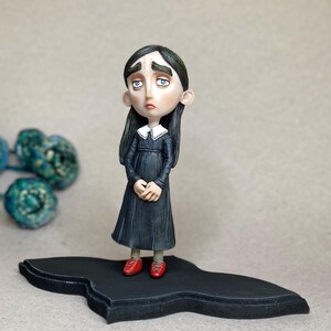 May include: A handcrafted figurine of a girl in a black dress with a white collar and red shoes. The doll has a melancholic expression and stands on a black, stylized base. The figure's long, dark hair frames a pale face.