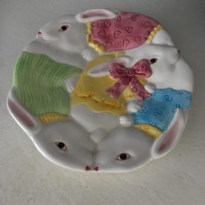 Op de afbeelding: Een wit keramiek bord in de vorm van een cirkel met vier konijnen erop. De konijnen dragen kleurrijke kleding en hebben roze oren. Het bord is versierd met een groen, geel, blauw en roze patroon.