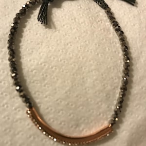 May include: A bracelet featuring small, dark grey faceted beads and a curved, copper-coloured bar. The beads are strung on a black cord, and the bar is embellished with small, clear crystals. The bracelet is tied with a black cord.