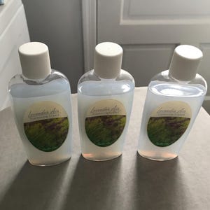 May include: Three clear glass bottles with white caps and labels that say "Lavender Air Simmering Liquid" with a picture of lavender flowers. The bottles are sitting on a white surface.