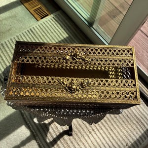 Puede incluir: Una cubierta rectangular de caja de pañuelos de metal dorado. La cubierta presenta un diseño calado ornamentado con detalles florales. La caja descansa sobre una pequeña mesa con un mantelito de encaje blanco. La estética general es vintage y decorativa.
