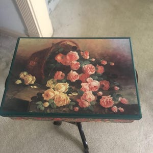 May include: A small wooden table with a floral design on the top. The table has a dark brown finish and a green border around the top. The floral design features a basket overflowing with pink and yellow roses.