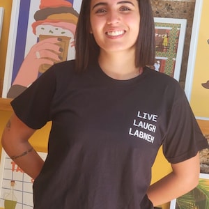 May include: A woman wearing a black t-shirt with the text "LIVE LAUGH LABNEH" printed on it. She is standing in front of a wall with various framed pictures.