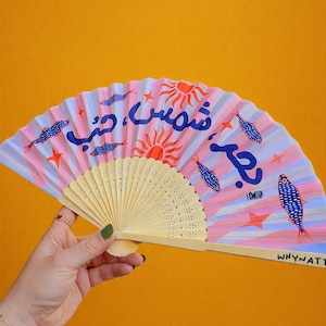 May include: A hand fan with a cream-colored handle and a colorful, patterned fan. The fan features a pink, blue, and white striped background with illustrations of fish, suns, and text. The text "WHYATT" is visible.
