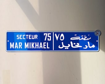 Puede incluir: Una señal de calle azul con texto blanco. La señal dice "Secteur 75 VO Mar Mikhael" en inglés y "مار مخايل  شعبة ٧٥" en árabe.