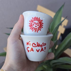 Puede incluir: Dos tazas de café de cerámica blanca apiladas, sostenidas en una mano. La taza superior tiene un diseño de sol rojo. La taza inferior tiene texto rojo en árabe y francés que dice "Hi, ça va?" Las tazas son pequeñas y parecen ser para espresso.