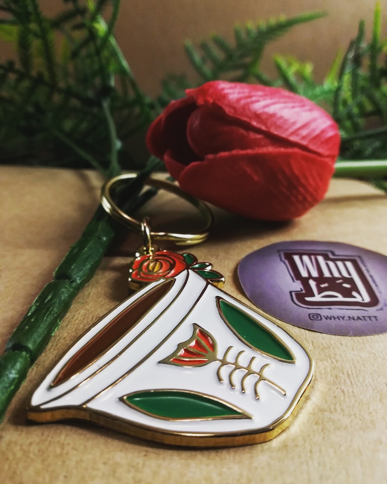 May include: Gold-tone metal keychain featuring a white coffee cup with a red rose and green leaves. The cup has a brown rim and a red rose with a green stem inside. The keychain is attached to a gold-tone ring.