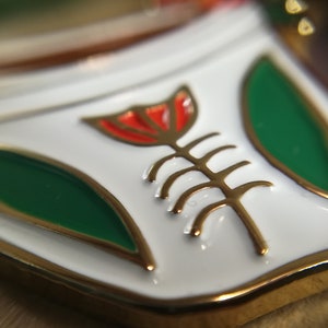 May include: A white enamel pin with a gold outline and a red flower with a gold stem and leaves. The flower is stylized and has a geometric design.