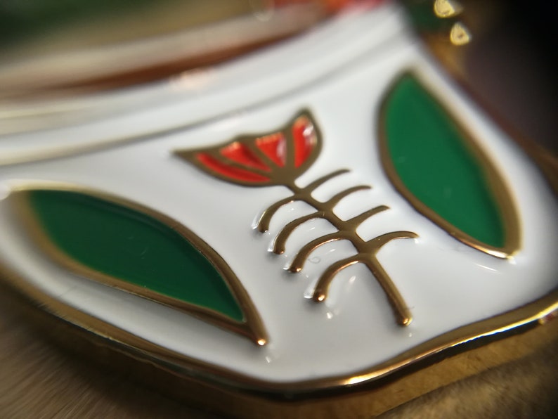 May include: A white enamel pin with a gold outline and a red flower with a gold stem and leaves. The flower is stylized and has a geometric design.