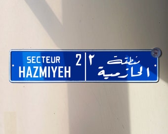 Puede incluir: Un letrero de calle rectangular azul con texto blanco. Muestra "SECTEUR HAZMIYEH" a la izquierda y escritura árabe a la derecha, con el número "2" en el centro. El sitio web "www.whynatt.com" está en la parte inferior.
